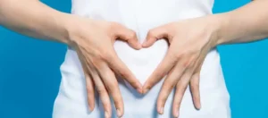 The image shows a person wearing a white shirt with their hands positioned in front of their abdomen to form the shape of a heart. The background is blue. This image evokes a sense of care, love, or attention toward the abdominal area—possibly indicating themes related to health, pregnancy, or well-being. Nice symbol for wellness! 