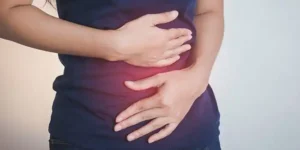 The image shows a person wearing a dark blue shirt, holding their stomach with both hands. The person’s left hand is placed on the upper abdomen, while the right hand is placed on the lower abdomen. There’s a red glow around the lower abdomen area, suggesting pain or discomfort in that region. This visual likely represents abdominal pain, which could indicate conditions like indigestion, cramps, or more serious gastrointestinal issues. No text or math in the image. 