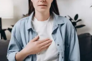 The image shows a person who appears to be placing their hand on their chest, suggesting they might be experiencing some discomfort or pain in that area. The gesture commonly indicates feelings like chest pain, heartburn, or emotional distress. The individual in the photo has a neutral to mildly concerned expression, which could hint at them trying to acknowledge or cope with their discomfort. The setting suggests it might be at home or in a relaxed environment, based on the casual clothing and background elements. This imagery is often associated with health-related contexts, particularly focusing on chest discomfort, breathing issues, or emotional sensitivity.