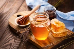 You Uploaded image Copilot The image shows a jar of honey placed on a wooden board. The jar is tied with a piece of twine around its neck. Next to the jar, there’s a honey dipper resting in a small white dish, which also contains some honey. There are also pieces of honeycomb placed on the wooden board. In the background, there is another jar, possibly containing a different type of honey or syrup, and a blue cloth. Additionally, there are sprigs of lavender scattered around the board. The setting appears to be rustic and natural, emphasizing the organic and wholesome nature of the honey. 