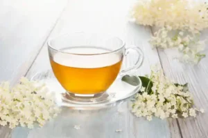 The image shows a clear glass cup filled with light amber-colored tea, placed on a matching glass saucer. Surrounding the cup are clusters of small, white flowers with green leaves, which appear to be elderflowers. The setting is on a light wooden surface, giving a fresh and natural feel to the scene. Elderflower tea is known for its health benefits and pleasant aroma.